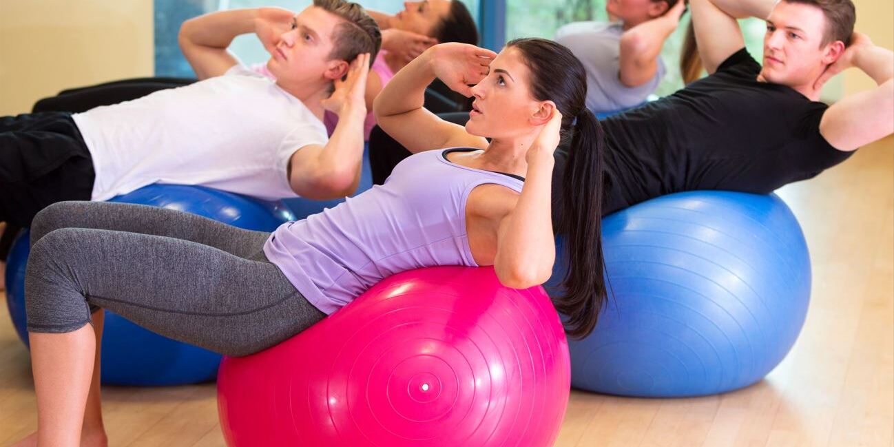 F&uuml;nf Personen machen Bauch&uuml;bungen auf Fitnessb&auml;llen in einem hellen Raum mit gro&szlig;en Fenstern.