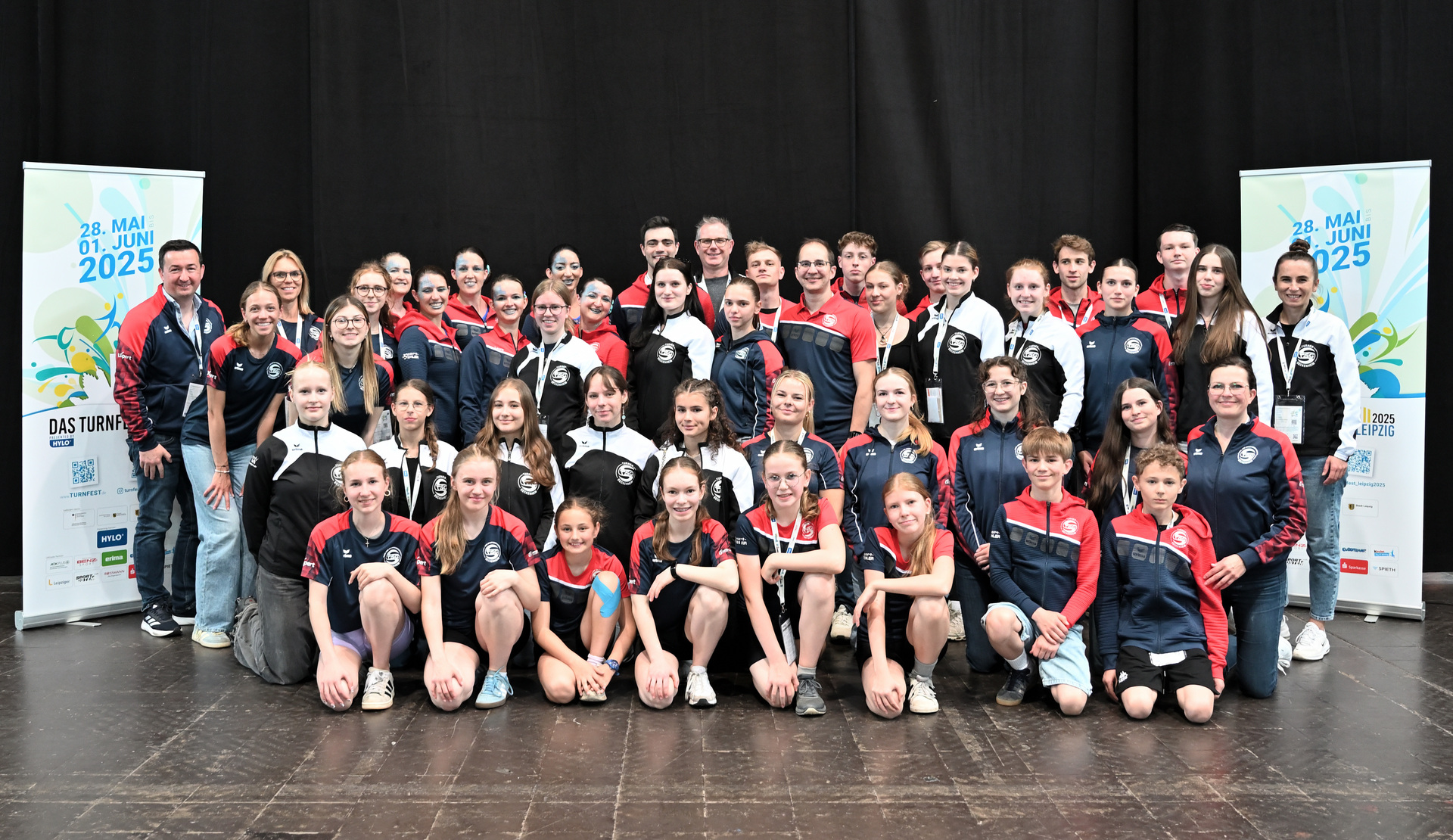 Gruppenfoto von Sportlern in Uniformen, stehend und sitzend vor Bannern f&uuml;r das Event in Leipzig 2025.