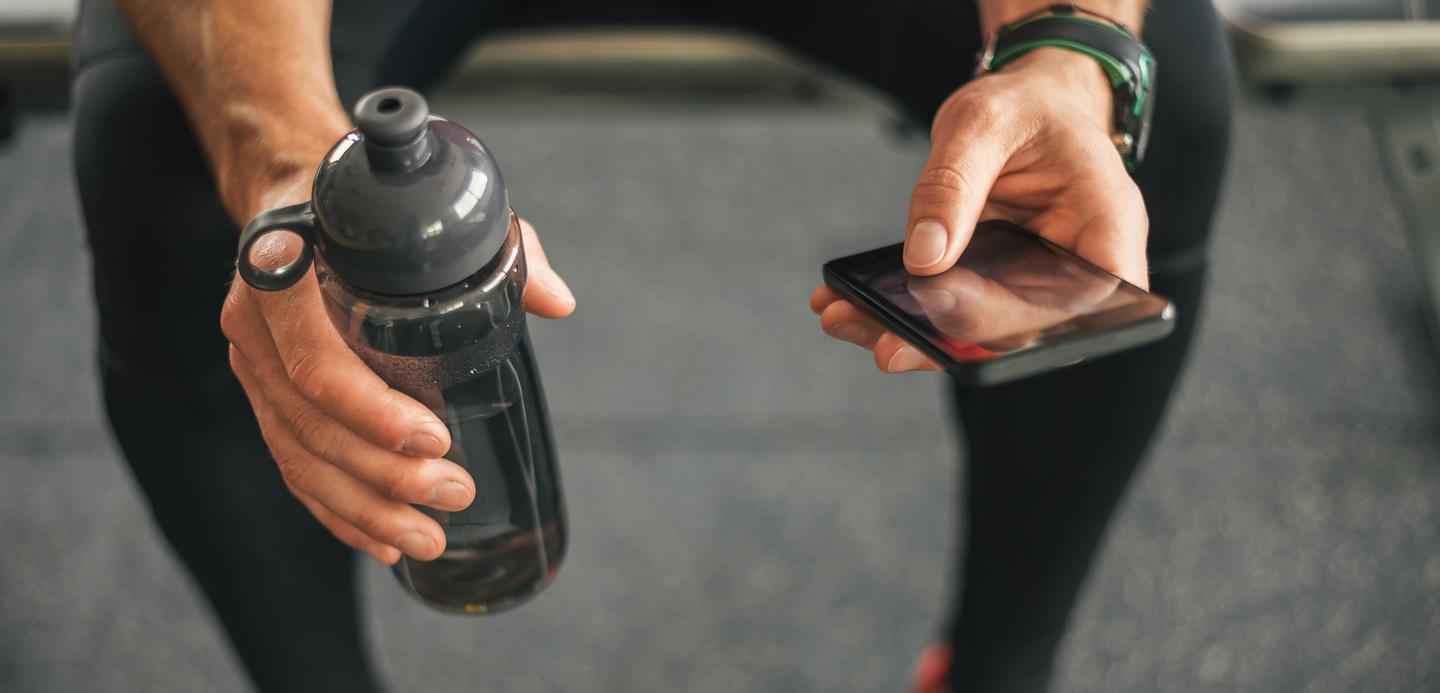 Mann sitzt auf einer Bank im Fitnessstudio, h&auml;lt eine Trinkflasche in der linken Hand und ein Smartphone in der rechten Hand.