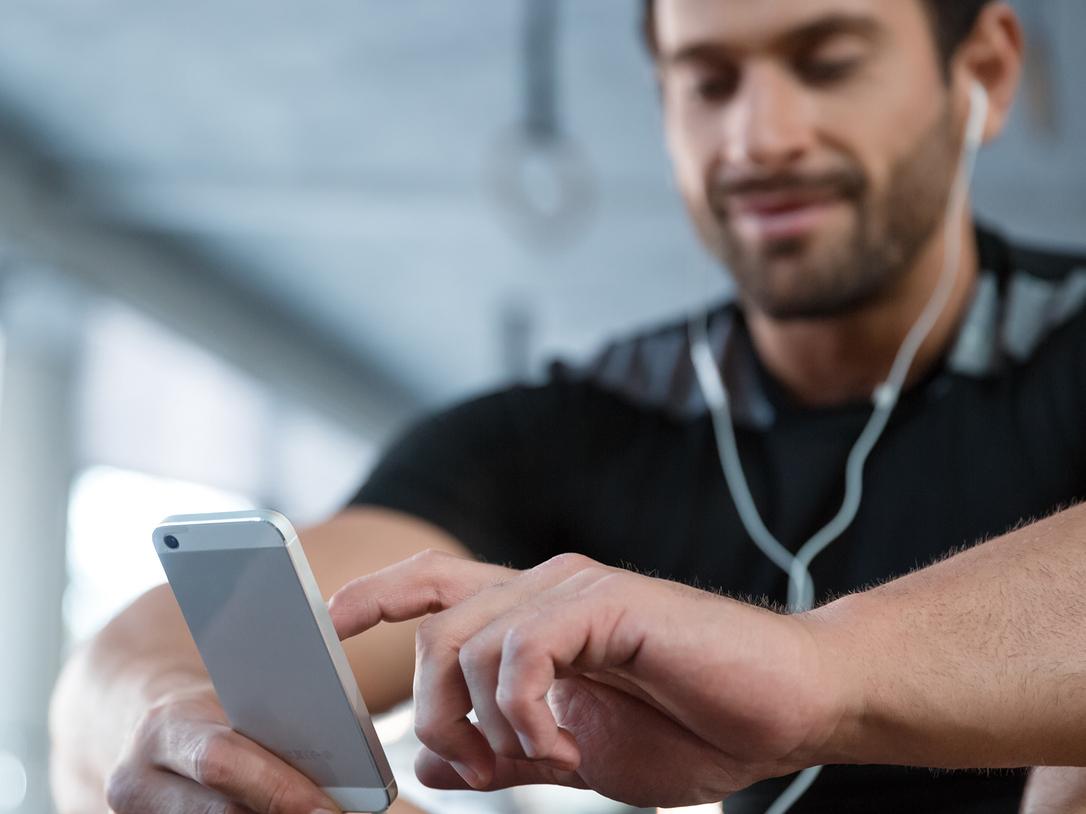 Mann mit Kopfh&ouml;rern sitzt und nutzt sein Smartphone in einem Fitnessstudio.
