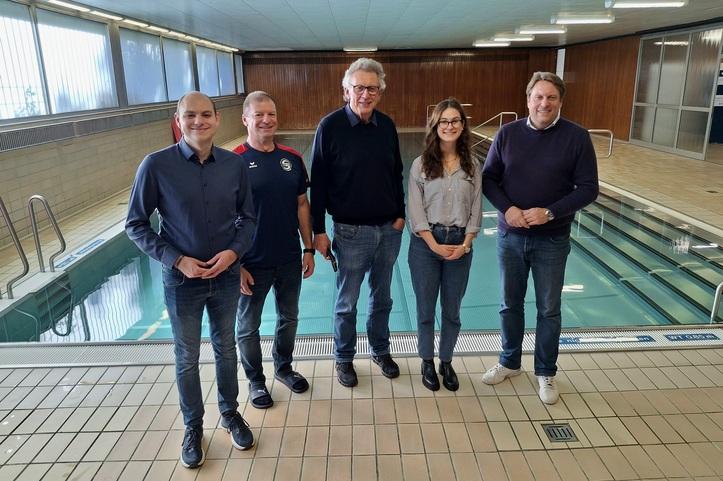 F&uuml;nf Personen stehen l&auml;chelnd am Rand eines Schwimmbeckens in einem hellen Schwimmbad.