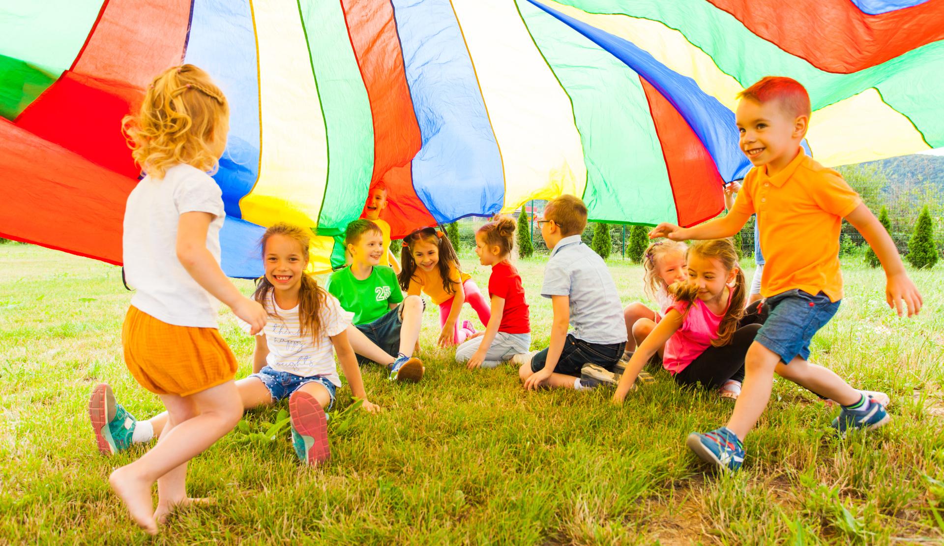 Eine Gruppe von Kindern spielt unter einem bunten Tuch auf einer Wiese und hat Spa&szlig; miteinander.