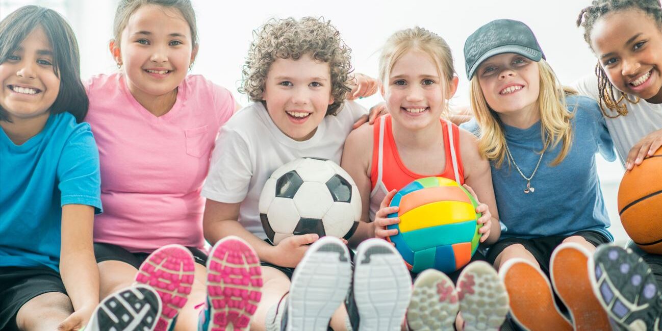 Sechs Kinder sitzen zusammen und halten Sportb&auml;lle, w&auml;hrend sie fr&ouml;hlich in die Kamera l&auml;cheln.