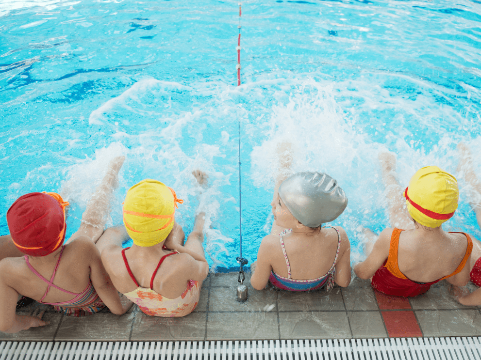 Gruppe von Kindern in Badekappen, die am Beckenrand sitzen und mit den F&uuml;&szlig;en im Wasser planschen.