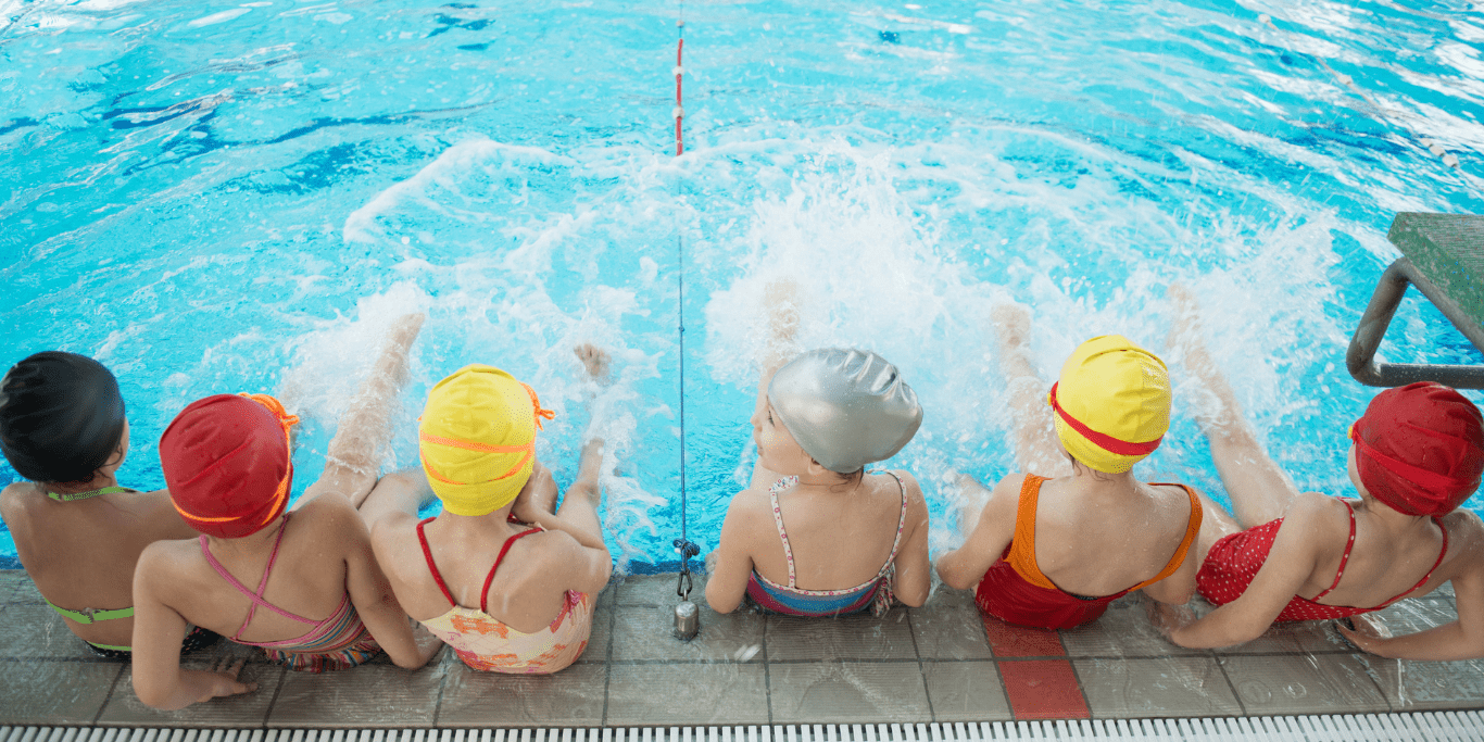 Gruppe von Kindern in Badekappen, die am Beckenrand sitzen und mit den F&uuml;&szlig;en im Wasser planschen.