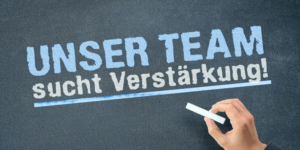 H&auml;nded mit Kreide schreibt &bdquo;UNSER TEAM sucht Verst&auml;rkung!&ldquo; auf eine dunkle, matte Tafeloberfl&auml;che.