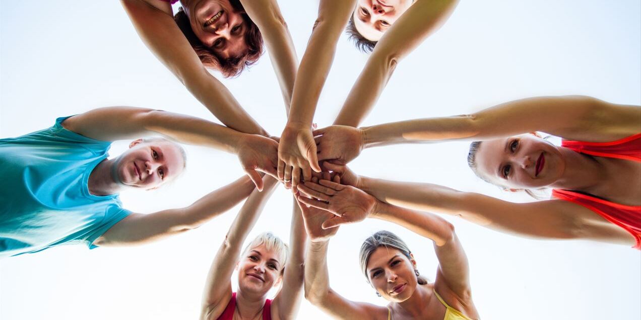 F&uuml;nf Frauen in bunten Sporttops formen einen Kreis mit erhobenen H&auml;nden, fotografiert von unten gegen einen hellen Himmel.