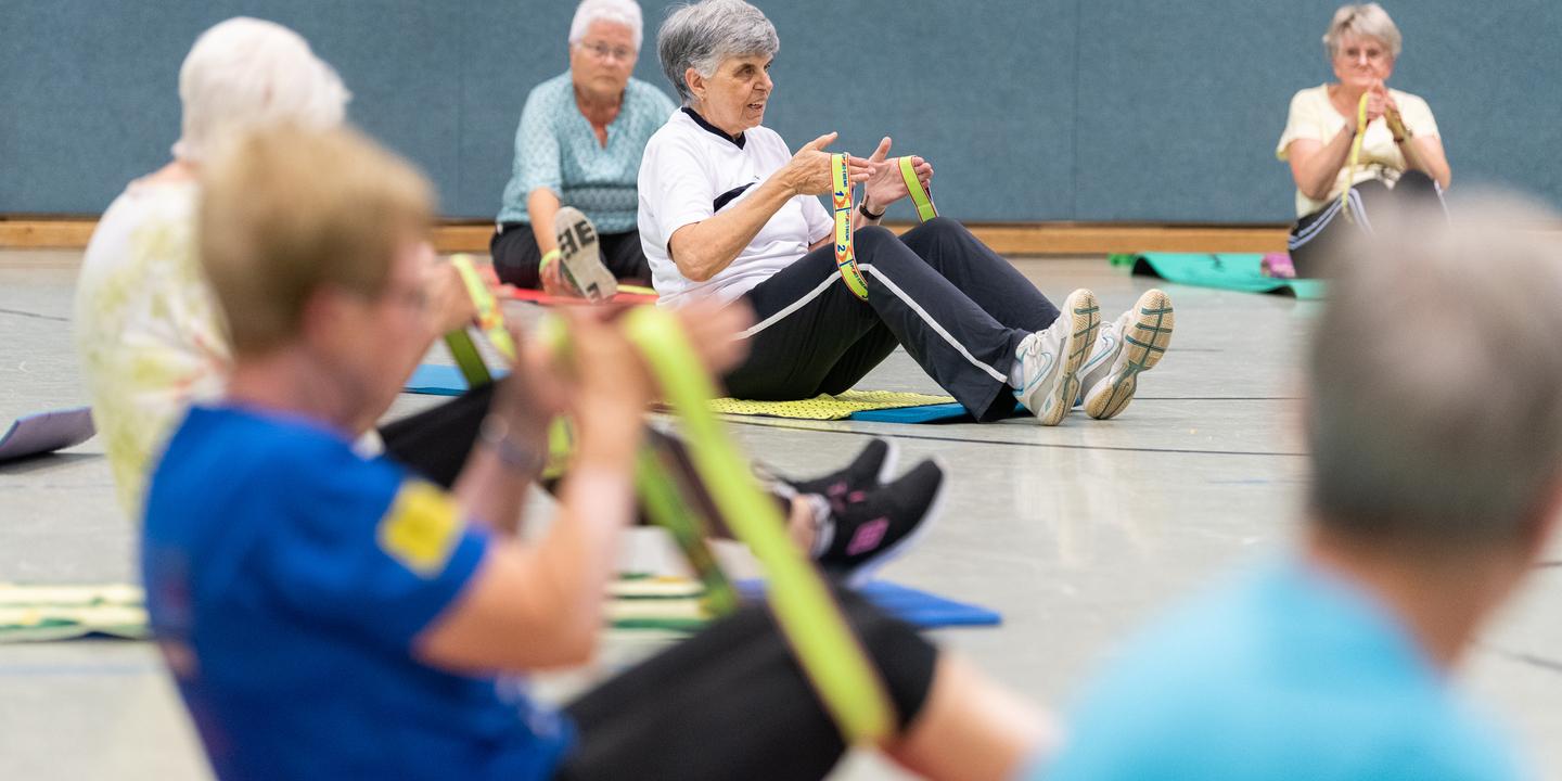 Senioren nehmen an einer Fitnessübung mit Fitnessbändern in einer Sporthalle teil, sitzend auf Fitnessmatten.