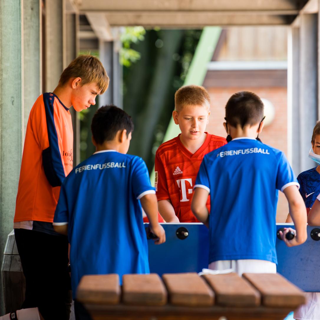 Sechs Kinder spielen gemeinsam mit einem Tischkicker unter einem überdachten Bereich.