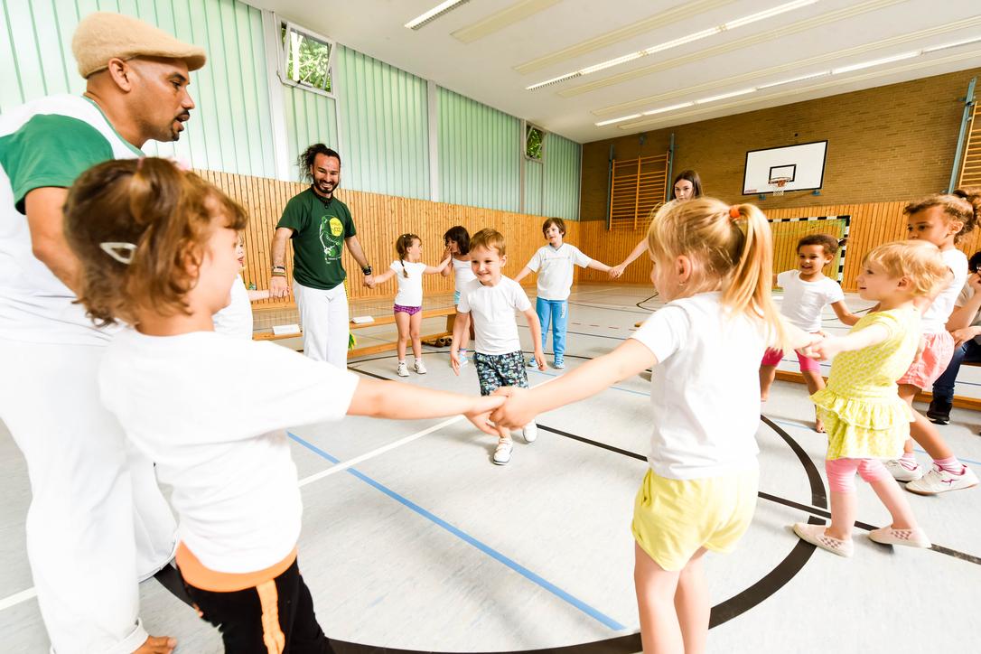 Gruppe von Kindern in T-Shirts und Erwachsenen in einer Turnhalle, die gemeinsam einen Kreis bilden und spielen.