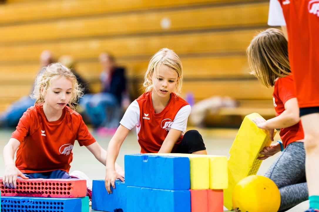 Drei Mädchen im roten Shirt bauen mit bunten Schaumstoffbausteinen in einer Turnhalle.