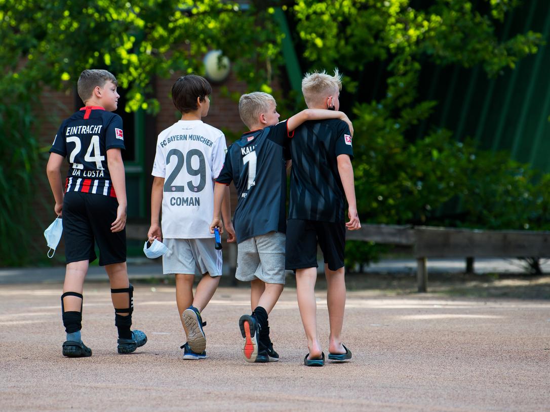 Vier Kinder in Fußballtrikots gehen Arm in Arm über einen Platz, umgeben von Bäumen und einer ruhigen Atmosphäre.