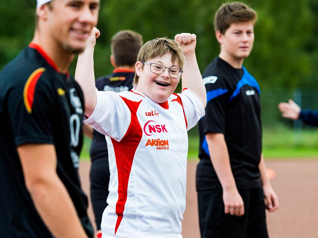 Ein junger Spieler in einem weißen Trikot hebt die Arme jubelnd, umgeben von weiteren Fußballspielern auf dem Platz.