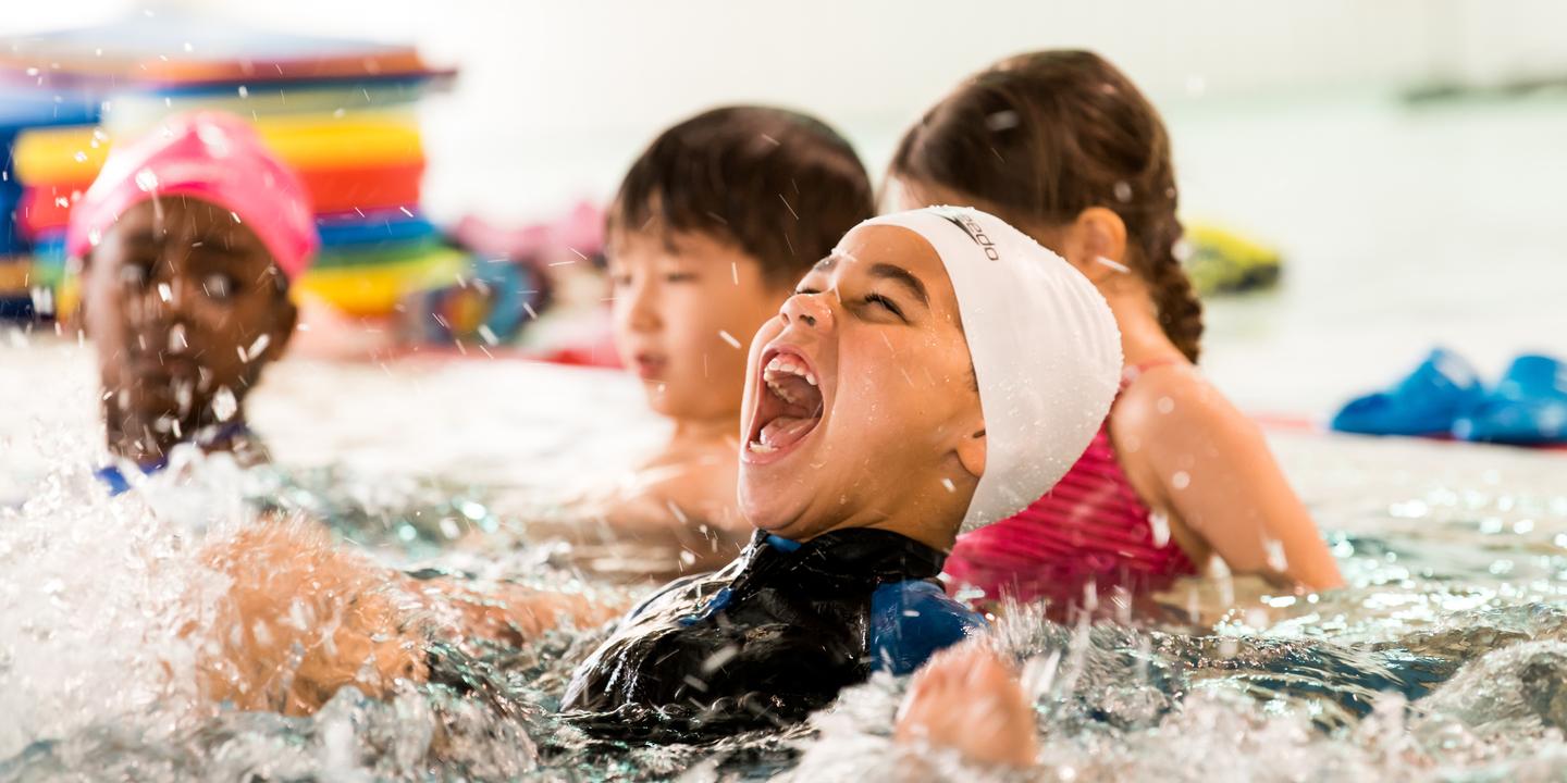 Vier Kinder spielen im Wasser eines Schwimmbads, ein Junge lacht laut und spritzt Wasser mit seinen Händen.