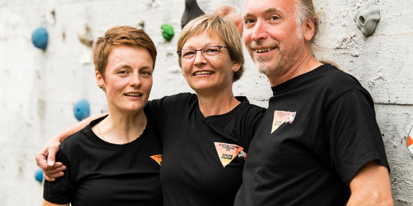 Drei Personen in schwarzen T-Shirts lächeln vor einer Kletterwand mit bunten Griffen.