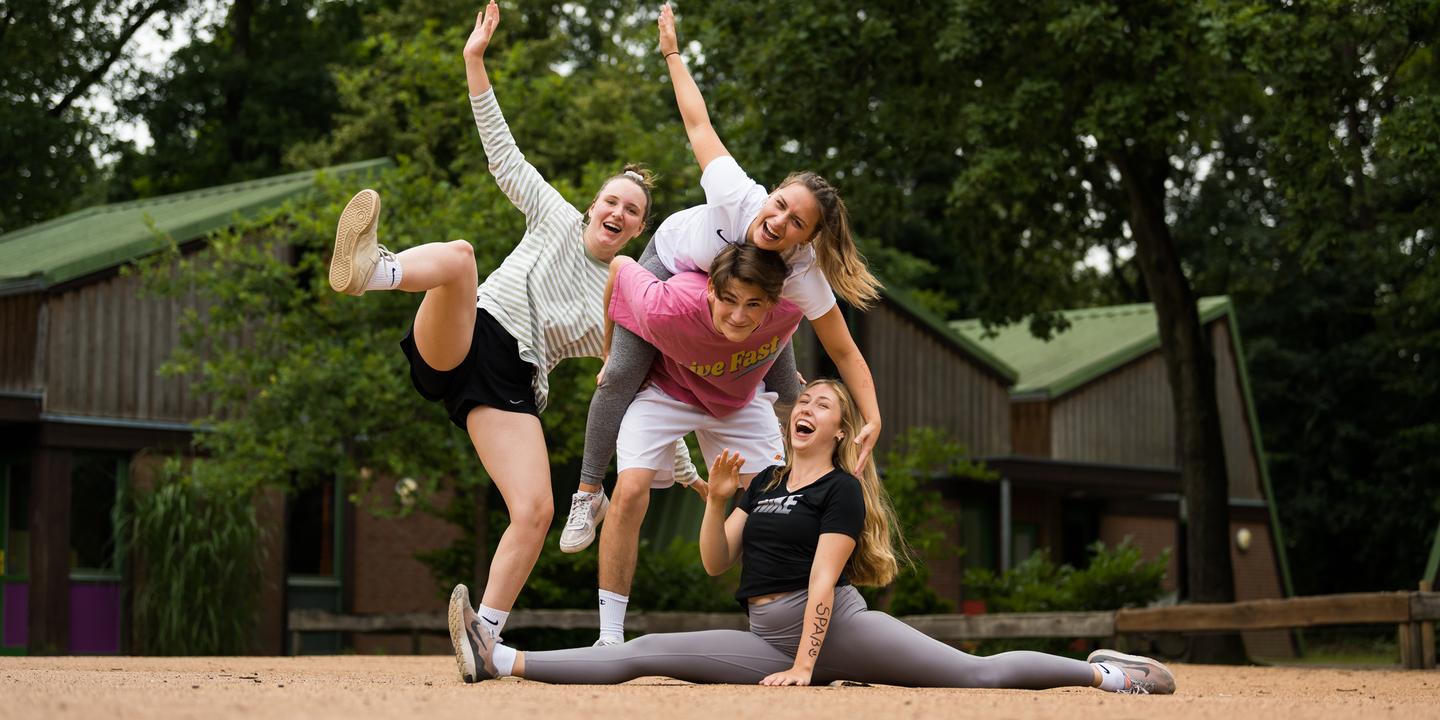 Vier junge Frauen posieren fröhlich auf einer sandigen Fläche, eine sitzt im Spagat, während die anderen sie umarmen.