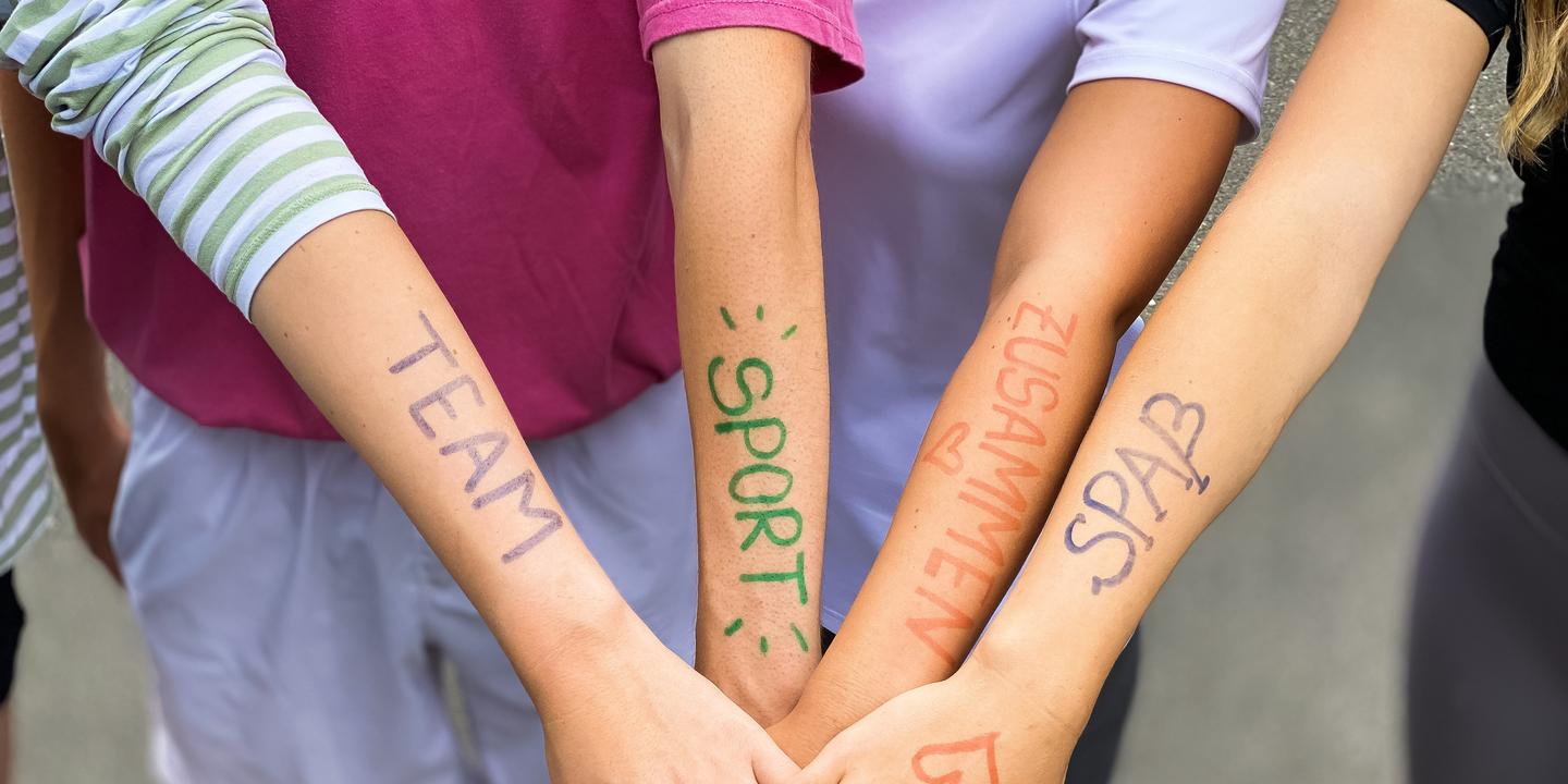 Vier Arme liegen übereinander, mit farbiger Schrift: „TEAM“, „SPORT“, „ZUSAMMEN“, „SPASS“ auf der Haut.