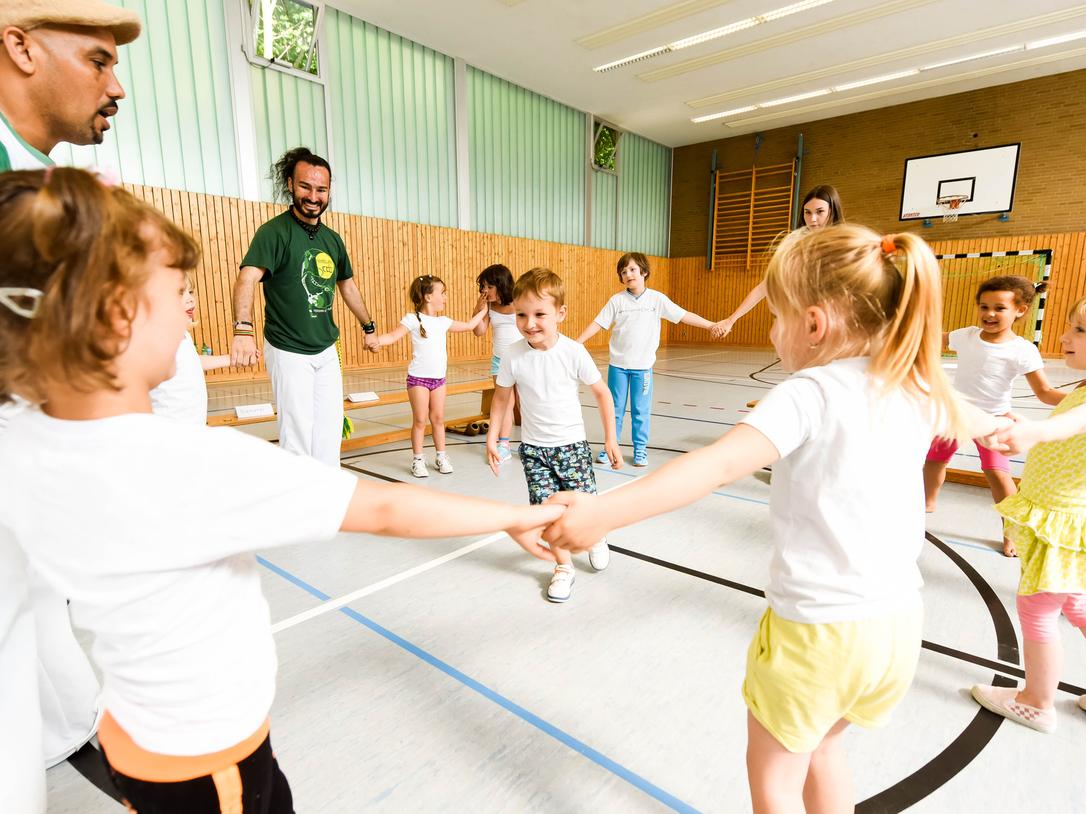 Gruppe von Kindern in T-Shirts und Erwachsenen in einer Turnhalle, die gemeinsam einen Kreis bilden und spielen.