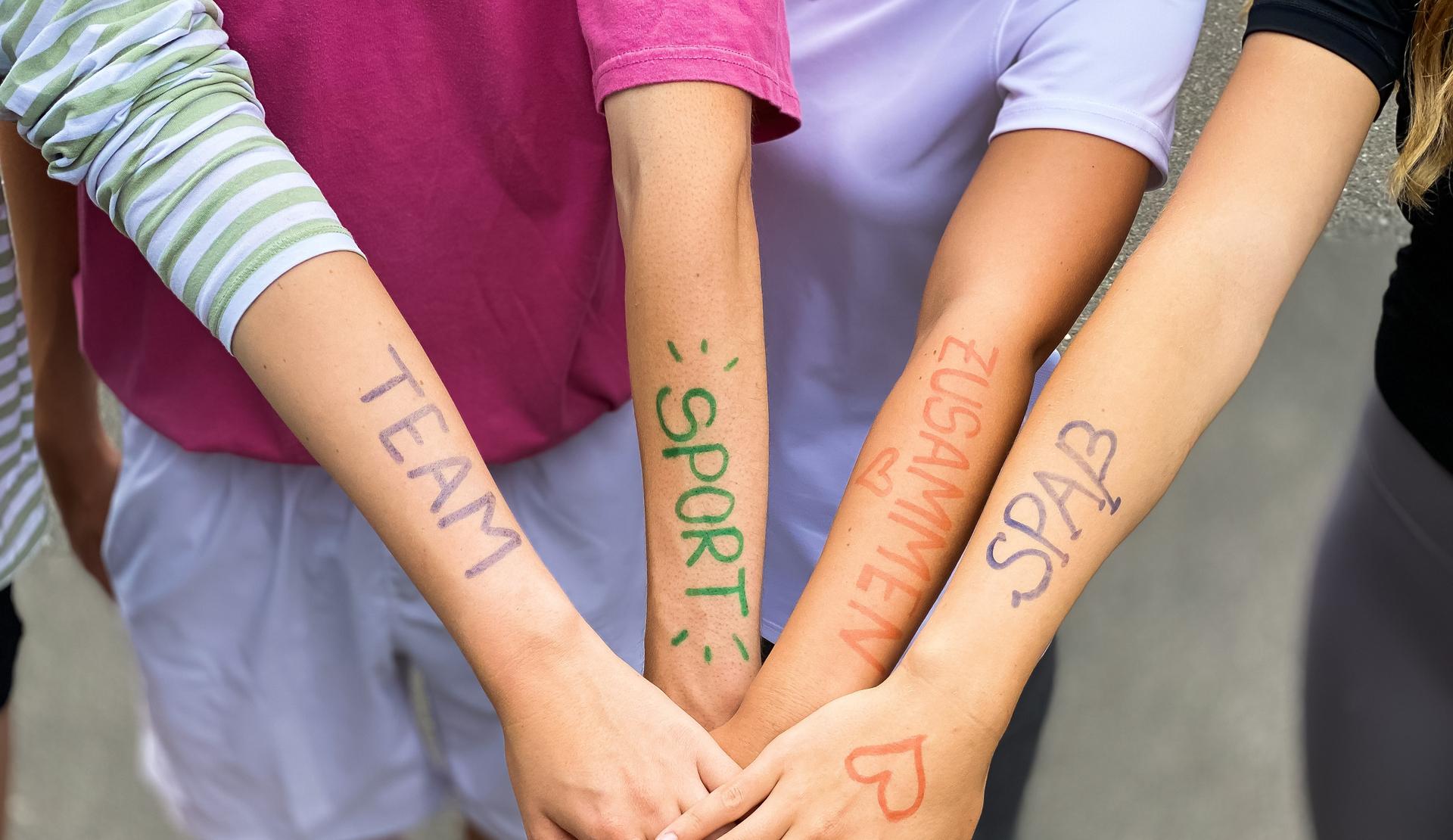 Vier Arme liegen übereinander, mit farbiger Schrift: „TEAM“, „SPORT“, „ZUSAMMEN“, „SPASS“ auf der Haut.