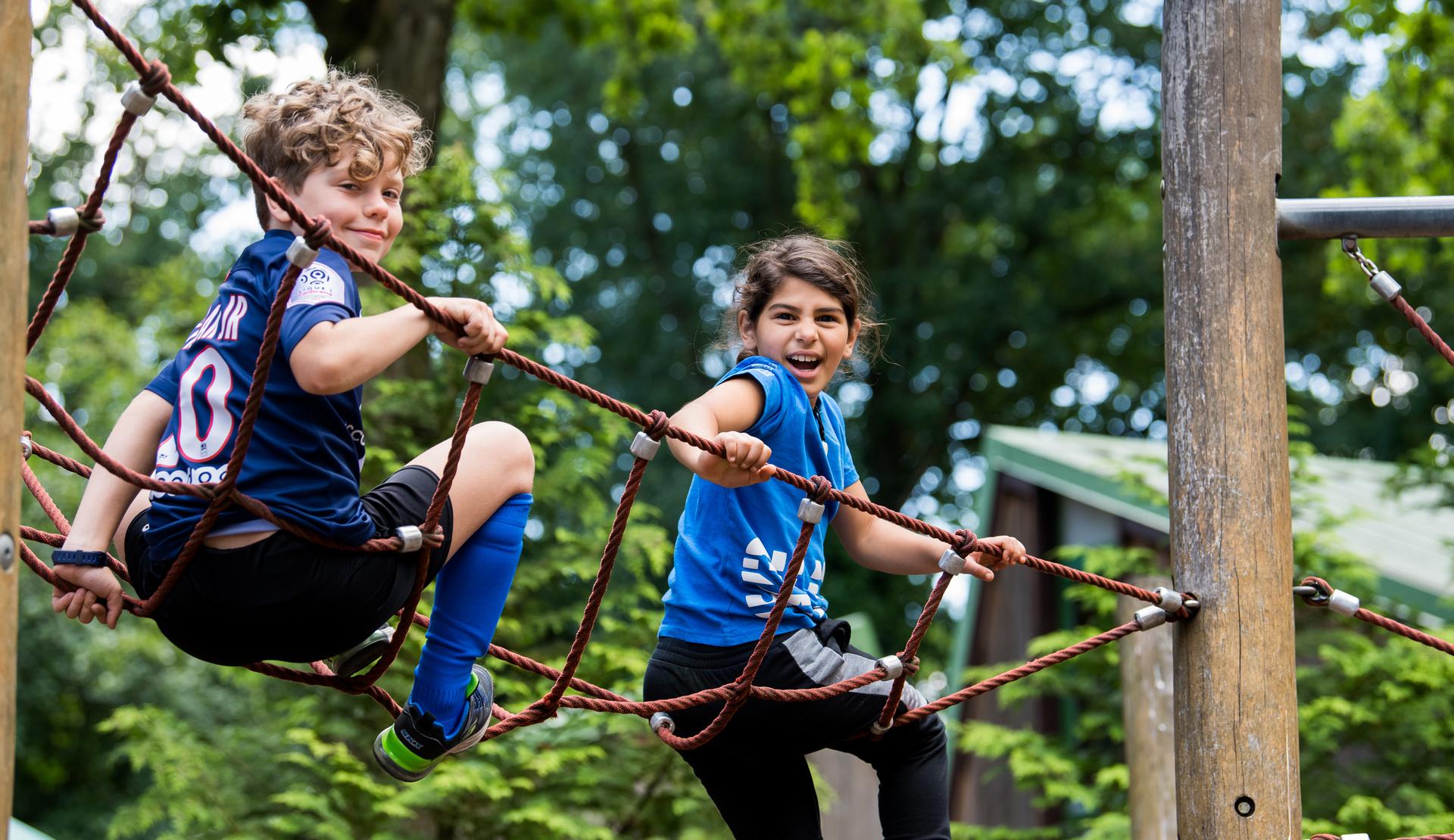 Zwei Kinder spielen auf einem Klettergerüst aus Seilen in einem Park, sie lachen und haben Spaß.