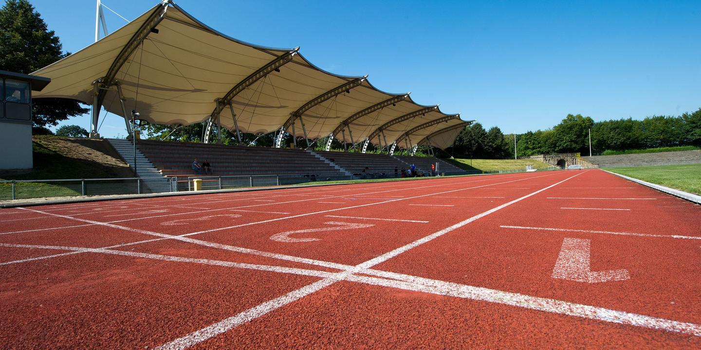 Nahaufnahme einer roten Laufbahn mit Startlinien und einem überdachten Zuschauerbereich im Hintergrund.