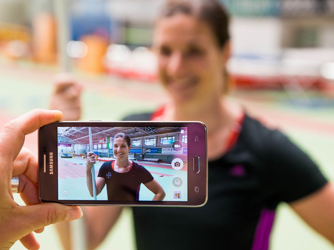 Hand hält ein Smartphone, das ein Porträt einer lächelnden Athletin mit Stab in einer Sporthalle zeigt.