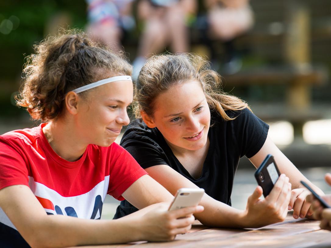 Zwei Mädchen sitzen an einem Tisch, schauen auf ihre Smartphones und lächeln dabei. Im Hintergrund sind weitere Personen sichtbar.