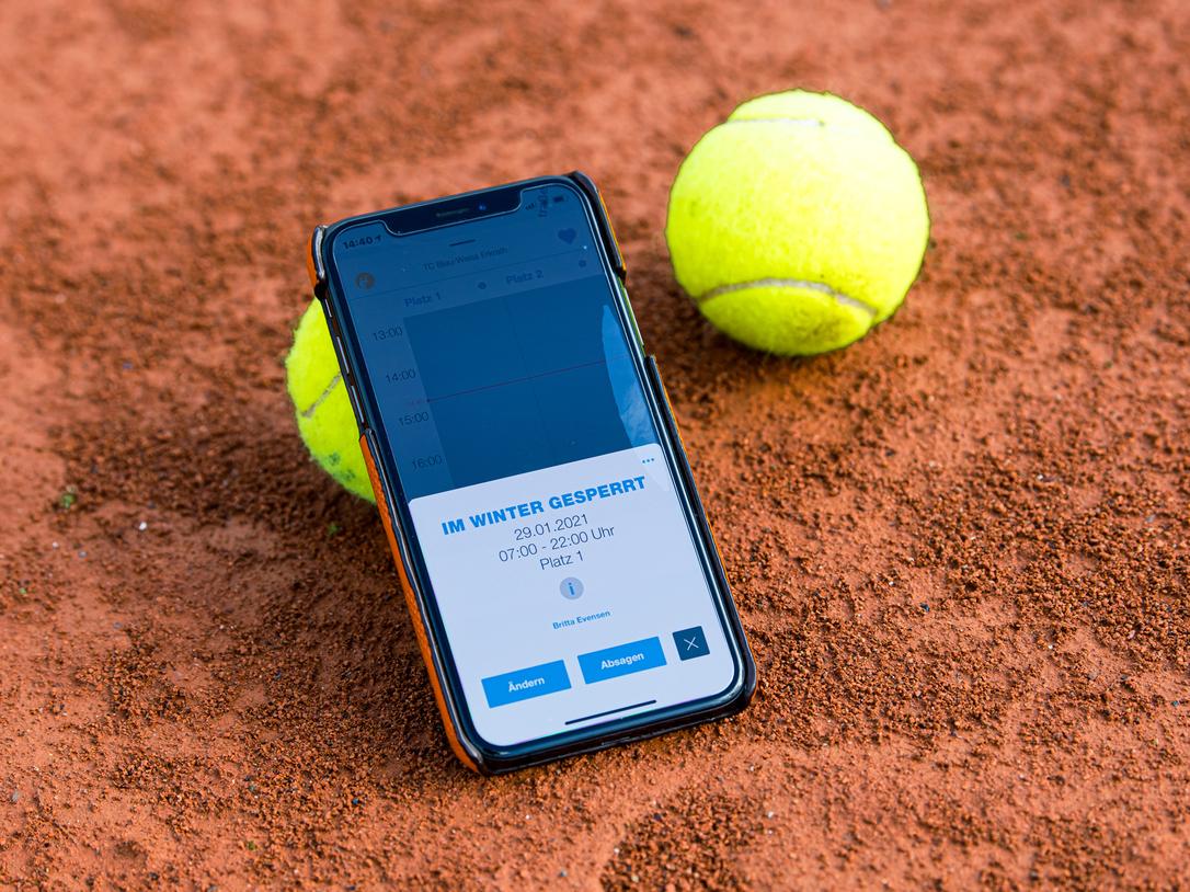 Smartphone mit Buchungsinfo liegt auf einem Tennisplatz, neben zwei gelben Tennisbällen auf rotem Sand.