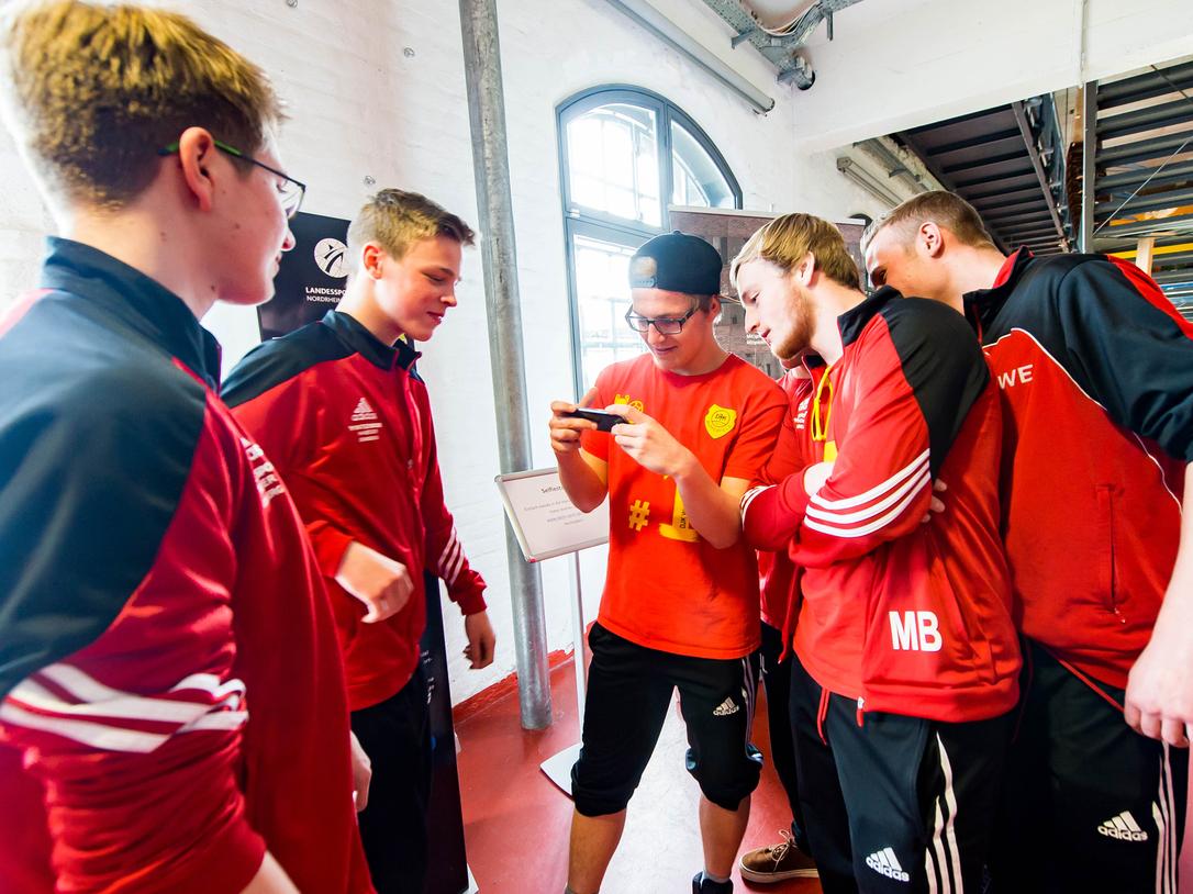 Sechs Personen in Sportbekleidung stehen zusammen und schauen auf ein Smartphone in einer hellen Umgebung.