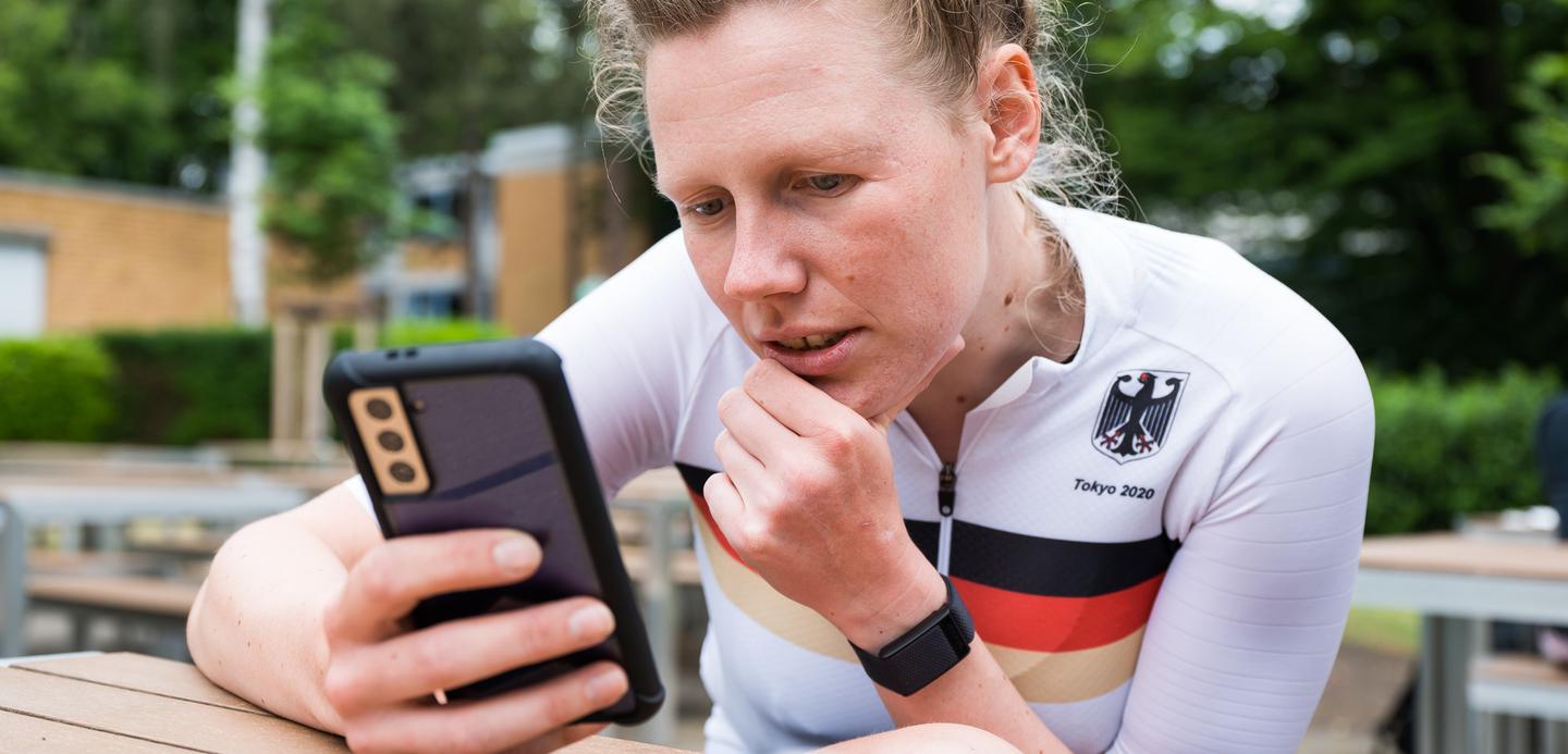 Athletin im weißen Trikot mit schwarzem und rotem Streifen schaut konzentriert auf ihr Smartphone in einer Außenanlage.