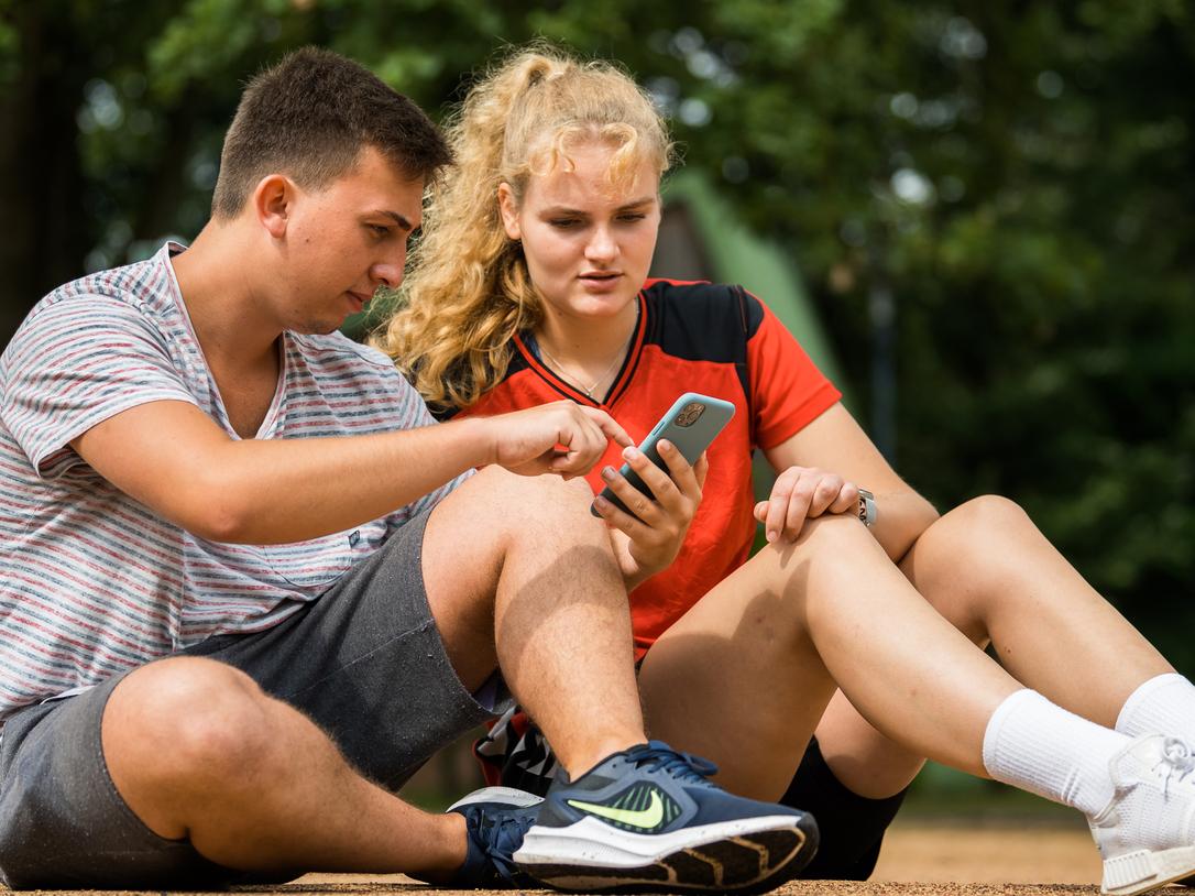 Zwei Jugendliche sitzen auf dem Boden und betrachten gemeinsam ein Smartphone. Ein Mädchen trägt ein rotes sportliches Oberteil.