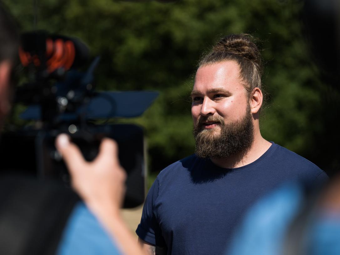 Mann mit Bart und Dutt steht vor einer Kamera und spricht, während ein weiteres Kamerateam im Hintergrund sichtbar ist.