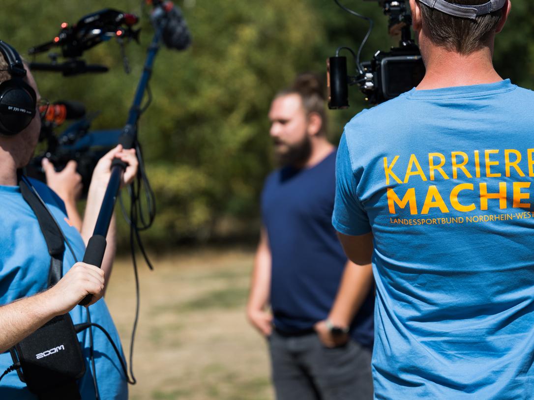 Drei Personen bei einer Videoaufnahme im Freien, einer trägt ein T-Shirt mit der Aufschrift „Karriere-Macher“.