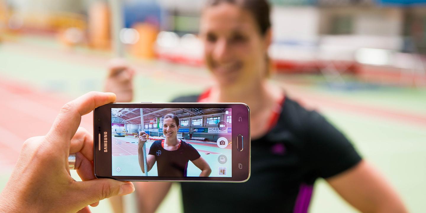 Hand hält ein Smartphone, das ein Porträt einer lächelnden Athletin mit Stab in einer Sporthalle zeigt.