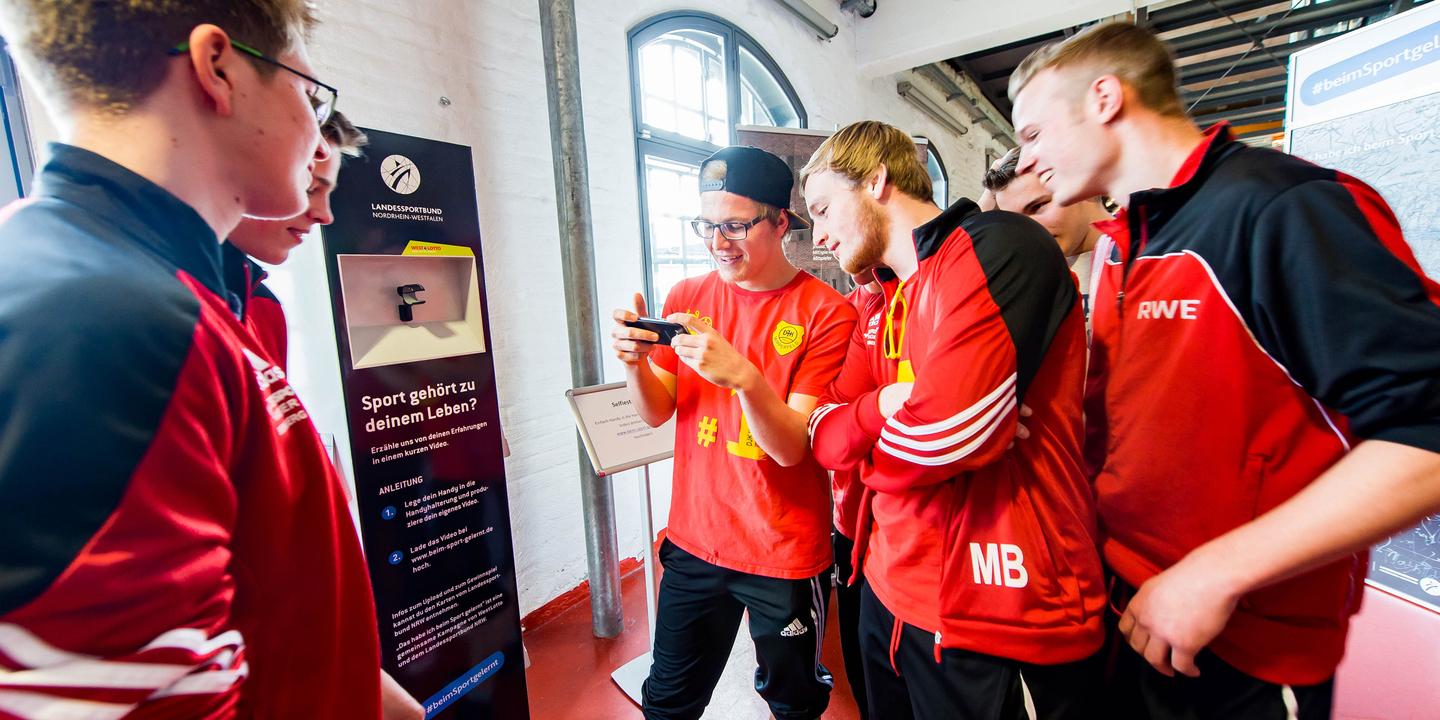 Gruppe junger Sportler in roten Trikots schaut auf ein Smartphone und diskutiert an einem Informationsstand.