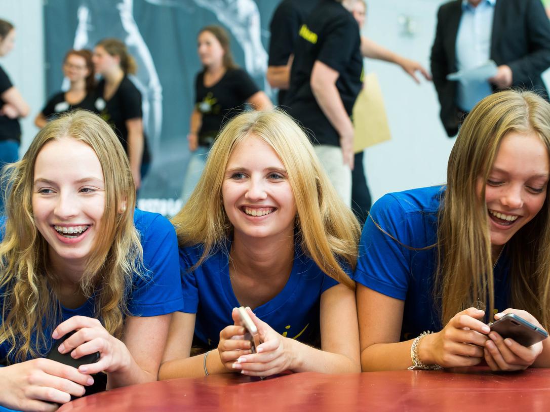 Drei lachende Jugendliche in blauen T-Shirts liegen auf einem Tisch und schauen auf ihre Handys. Hintergrund mit weiteren Personen.