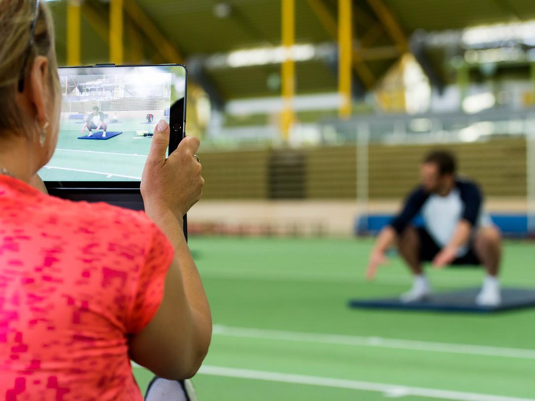 Eine Person filmt einen Mann, der auf einer Matte in der Squat-Position trainiert, in einem Sportzentrum.