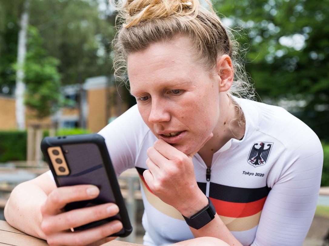 Athletin im weißen Trikot mit schwarzem und rotem Streifen schaut konzentriert auf ihr Smartphone in einer Außenanlage.