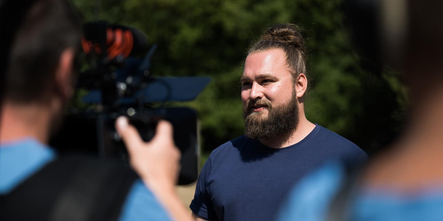 Mann mit Bart und Dutt steht vor einer Kamera und spricht, während ein weiteres Kamerateam im Hintergrund sichtbar ist.