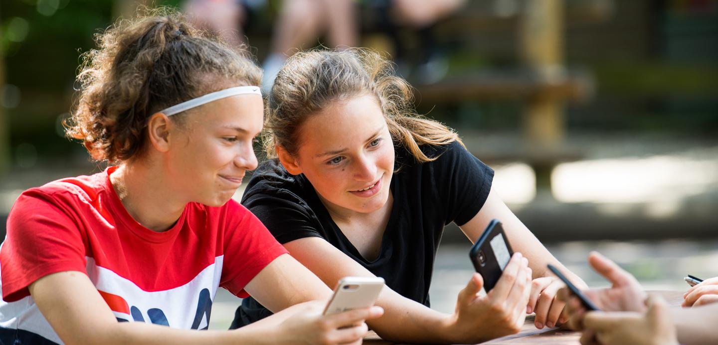 Zwei Mädchen sitzen an einem Tisch, schauen auf ihre Smartphones und lächeln dabei. Im Hintergrund sind weitere Personen sichtbar.