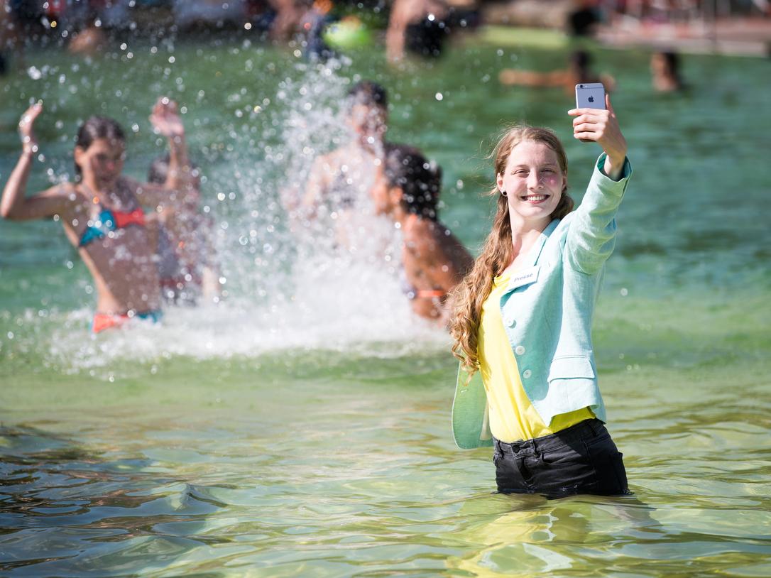 Lächelnde Frau im Wasser, die ein Selfie macht, während Kinder im Hintergrund spielen und spritzen.