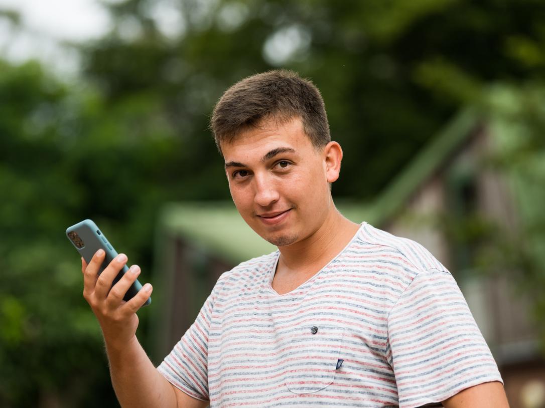 Junger Mann mit kurzen Haaren hält ein Smartphone in der Hand und schaut lächelnd in die Kamera, Hintergrund unscharf.