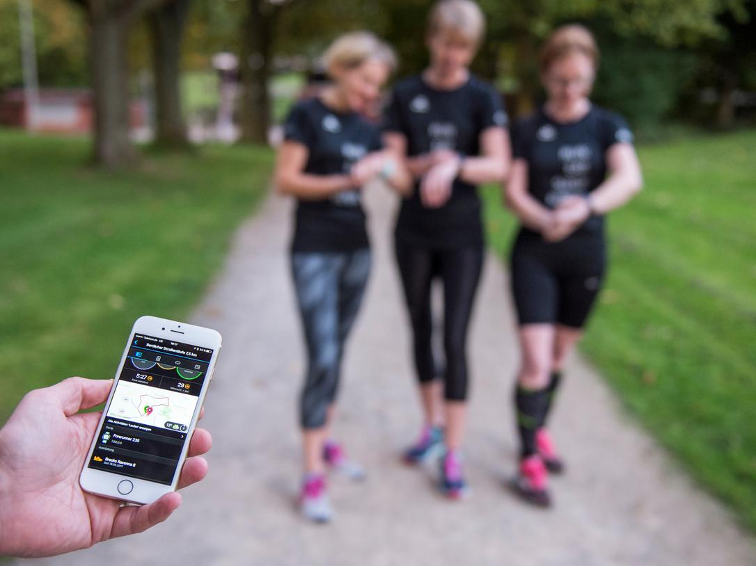 Ein Mann hält ein Smartphone mit Fitness-App, während drei Frauen in Sportkleidung auf einem Spazierweg stehen.