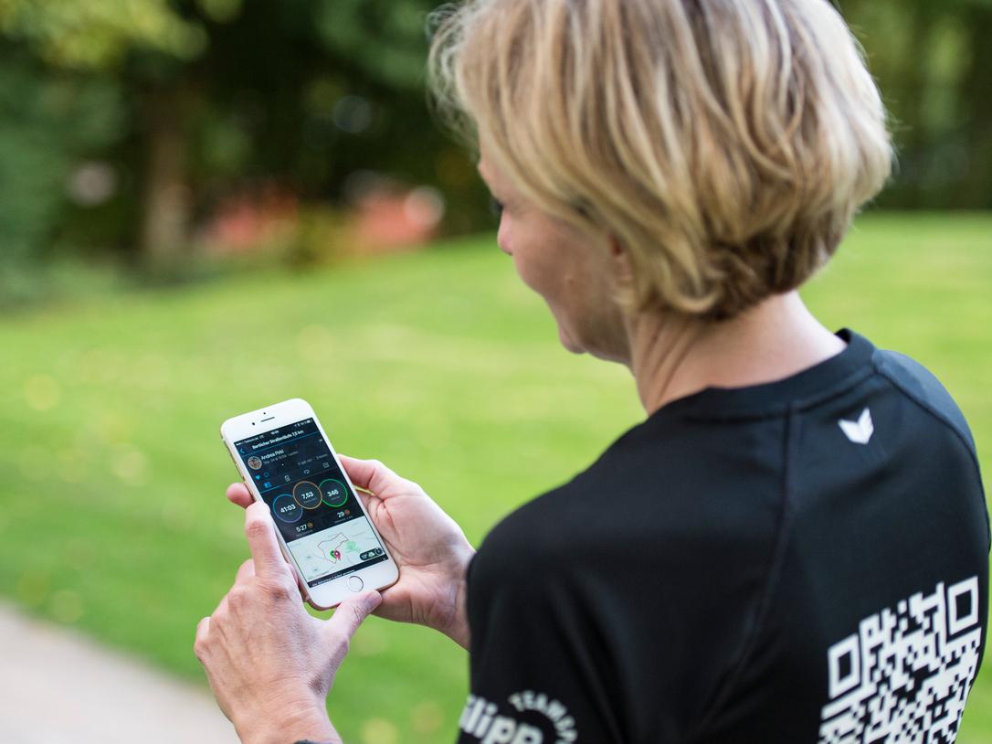 Frau mit kurzen Haaren schaut auf ein Smartphone, während sie auf einem Weg in einem Park steht.