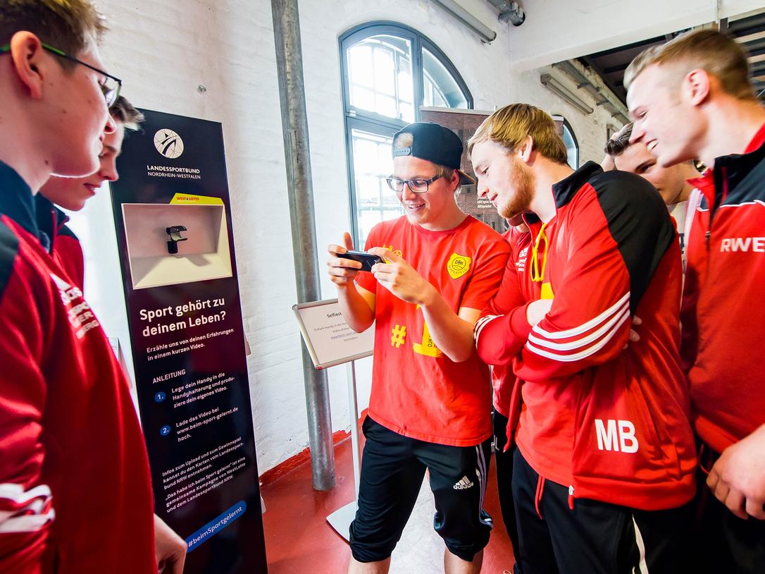 Gruppe junger Sportler in roten Trikots schaut auf ein Smartphone und diskutiert an einem Informationsstand.