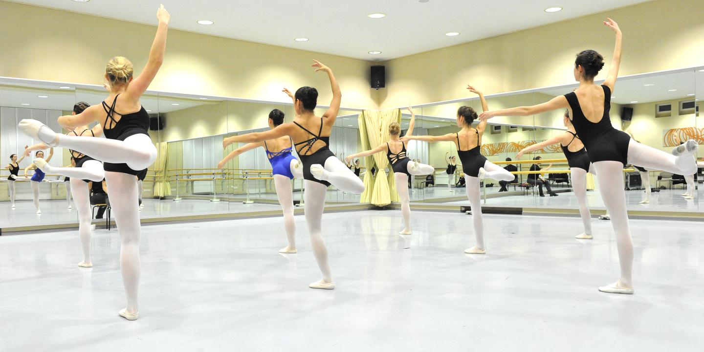 Gruppe von Balletttänzerinnen in Schwarz und Blau übt in einem Spiegelraum mit Holzballettstangen.