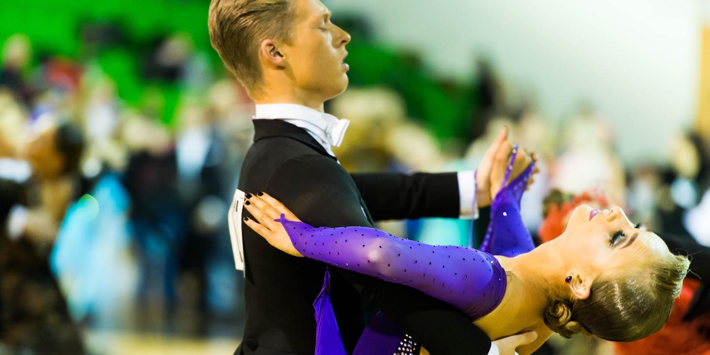 Tanzpaar in eleganter Haltung auf der Tanzfläche, die Frau trägt ein glitzerndes lila Kleid und lächelt.