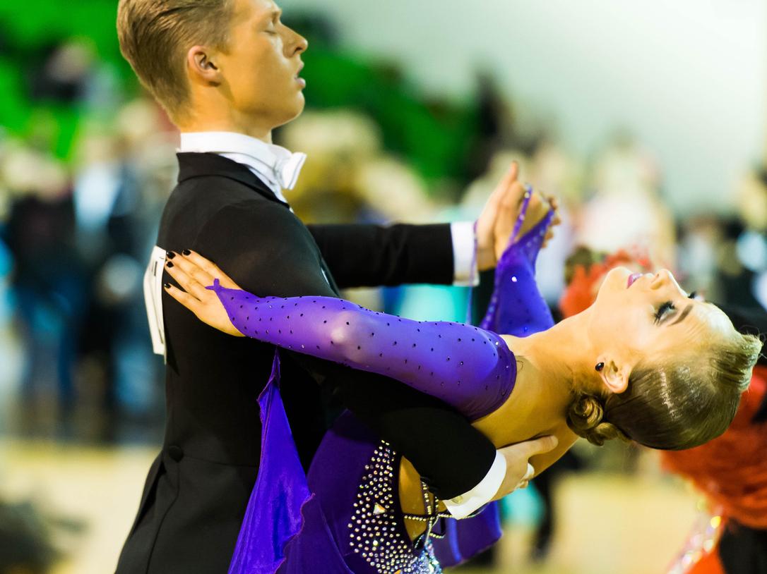 Tanzpaar in eleganter Haltung auf der Tanzfläche, die Frau trägt ein glitzerndes lila Kleid und lächelt.