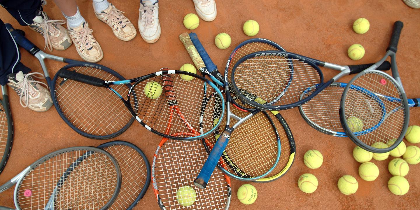 Ansammlung von Tennisschlägern und Bällen auf einem Sandplatz, umgeben von Füßen in Sportschuhen.