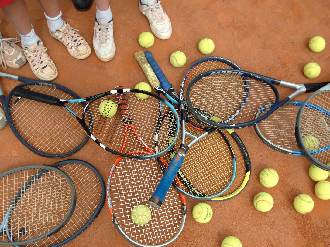Ansammlung von Tennisschlägern und Bällen auf einem Sandplatz, umgeben von Füßen in Sportschuhen.